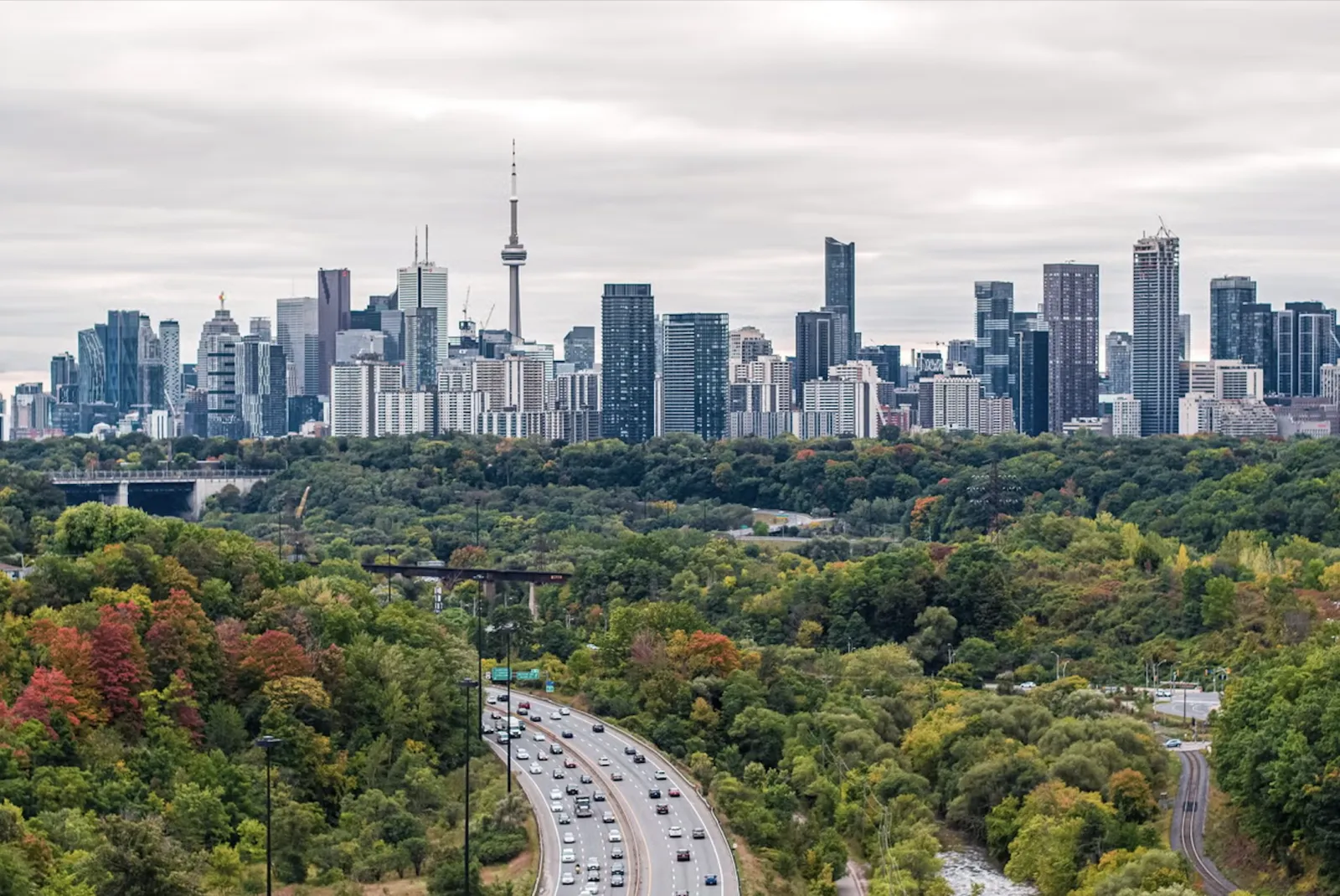 Toronto's downtown skyline and the CN Tower rising behind a band of autumn trees, with the Don Valley Parkway curving through the foreground.